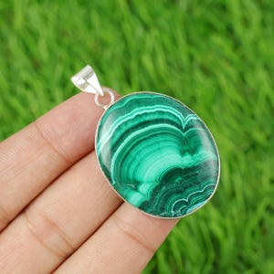 Puede incluir: Colgante ovalado de malaquita con una fianza de plata. El colgante presenta un color verde vibrante con patrones arremolinados. La piedra está engastada en un bisel de plata. El colgante mide aproximadamente 3,8 cm de largo.