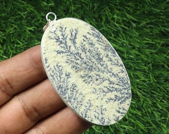 May include: Oval pendant with a silver-colored frame. The pendant features a cream-colored stone with a black, tree-like pattern. The pendant is held in a hand against a green background.