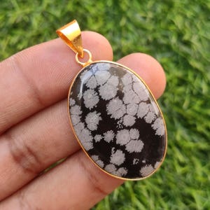 Puede incluir: Colgante ovalado con una piedra negra con patrones blancos en forma de copo de nieve, engastada en un marco de metal dorado. Tiene una fianza dorada para una cadena. El fondo es verde borroso.