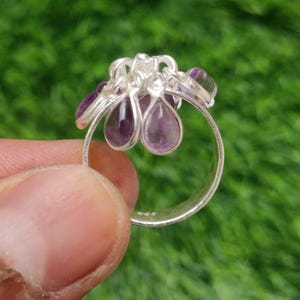 May include: A silver ring with a cluster of purple amethyst gemstones. The ring has a floral design with teardrop-shaped stones. The ring is held against a green background.