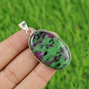 Puede incluir: Colgante ovalado con una piedra verde pulida con inclusiones rojas y negras, engastada en un marco de metal plateado. El colgante tiene un lazo para una cadena. Fondo verde borroso.