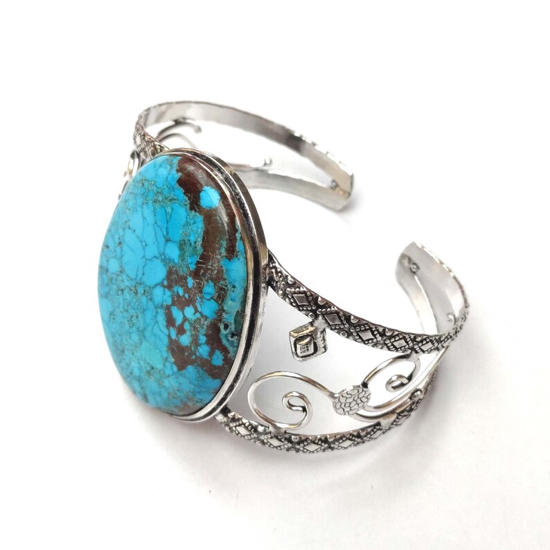 May include: A silver cuff bracelet featuring a large turquoise stone set in the centre. The bracelet has a delicate, ornate design with swirls and diamonds.