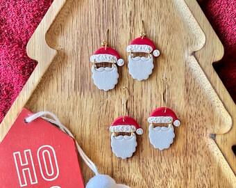May include: Four Santa Claus earrings with red hats, white beards, and gold hooks are displayed on a wooden tree-shaped tray. A red tag with the words "HO HO HO" is attached with a white string and a white pom-pom.