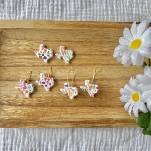 May include: A set of four floral Texas-shaped earrings. The earrings are made of white clay with a floral pattern and gold hoops. The earrings are displayed on a wooden tray.