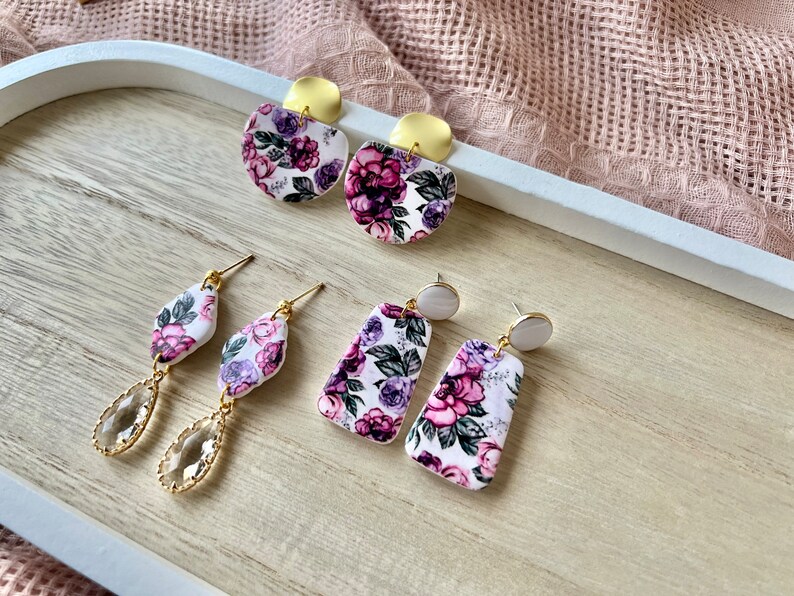 May include: A set of four floral patterned earrings with gold and white accents. The earrings are displayed on a wooden surface.