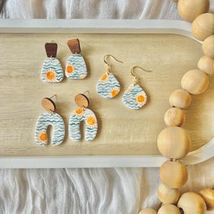 May include: A collection of handmade earrings with a beach theme. The earrings feature a blue and white wave pattern with a yellow sun design. Some earrings have wooden accents. The earrings are displayed on a wooden tray.
