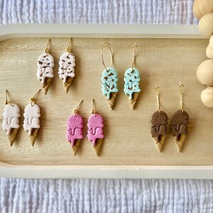 May include: Six pairs of ice cream cone earrings. The earrings are made of polymer clay and come in a variety of colors, including white, pink, blue, and brown. The earrings are all in the shape of a double-scoop ice cream cone with chocolate sprinkles.