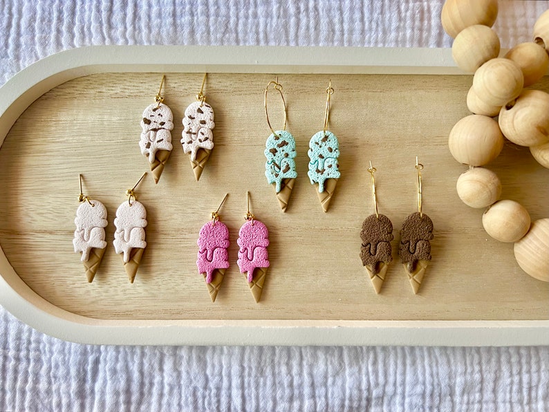 May include: Six pairs of ice cream cone earrings. The earrings are made of polymer clay and come in a variety of colors, including white, pink, blue, and brown. The earrings are all in the shape of a double-scoop ice cream cone with chocolate sprinkles.