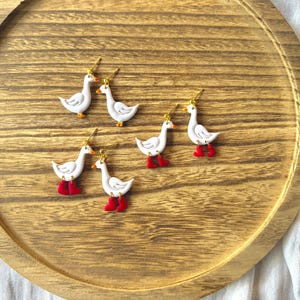 May include: Six white goose earrings with gold-colored hooks and red boots are arranged on a wooden tray. The geese have orange beaks and feet. The tray is round with a light brown finish, and the background is a white cloth.