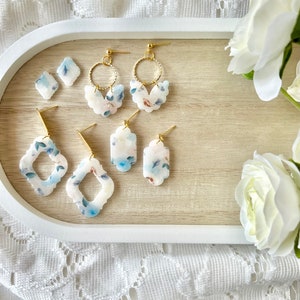 May include: A set of five pairs of earrings featuring floral patterns in shades of blue and white. The earrings are made of polymer clay and have gold-toned metal findings.