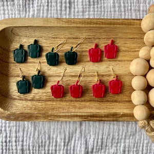 May include: Six pairs of earrings featuring red and green bell pepper shapes. The earrings are displayed on a wooden tray.