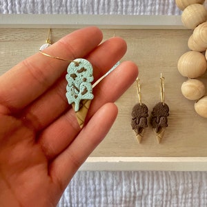 May include: A pair of gold hoop earrings with ice cream cone charms. One charm is mint green with brown sprinkles and the other is brown with white sprinkles.