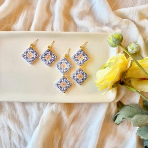 May include: Six pairs of square earrings with a blue and yellow floral design on a white background. The earrings have gold-colored hooks. The earrings are displayed on a white rectangular tray, with yellow flowers in the background.