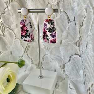 May include: A pair of white and purple floral earrings with gold accents. The earrings are rectangular in shape and feature a white background with a floral pattern in shades of purple, pink, and green. The earrings are hanging from a white post with a gold hook.