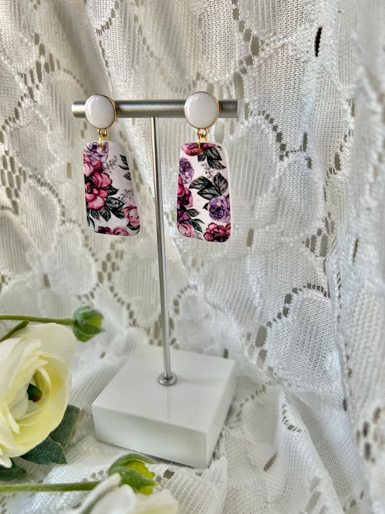 May include: A pair of white and purple floral earrings with gold accents. The earrings are rectangular in shape and feature a white background with a floral pattern in shades of purple, pink, and green. The earrings are hanging from a white post with a gold hook.