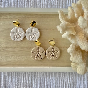 May include: Two pairs of earrings with a beach theme. The earrings feature a gold-colored, irregular-shaped top and a round, sand dollar-shaped bottom. One pair is white, and the other is tan. The earrings are displayed on a light wooden tray.