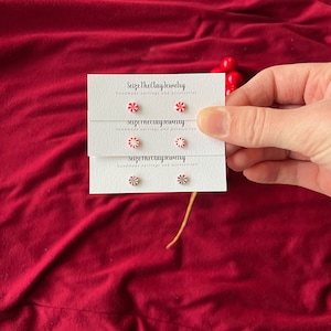 May include: Handmade stud earrings with festive designs are displayed on white cards. The earrings feature red and white peppermint candy and snowflake designs. The cards are held against a red fabric background. The text "Seize The Clay Jewelry" is visible.