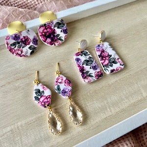 May include: Three pairs of floral earrings with gold accents. The first pair is a round shape with a white background and purple flowers. The second pair is a rectangular shape with a white background and purple flowers. The third pair is a teardrop shape with a clear crystal and a white background with purple flowers.