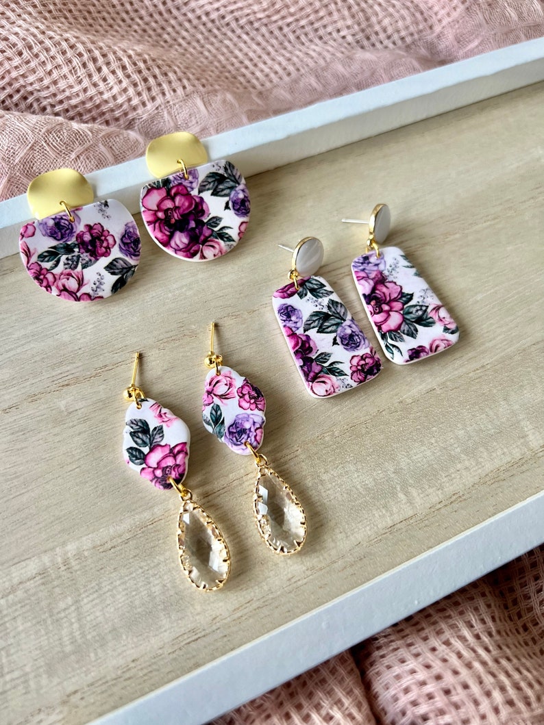 May include: Three pairs of floral earrings with gold accents. The first pair is a round shape with a white background and purple flowers. The second pair is a rectangular shape with a white background and purple flowers. The third pair is a teardrop shape with a clear crystal and a white background with purple flowers.