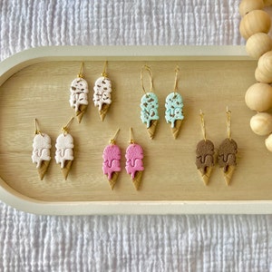 May include: A set of six pairs of ice cream cone earrings. The earrings are made of polymer clay and feature different ice cream flavors, including white, pink, mint green, and brown. The earrings are all gold-plated and have a dangling design.