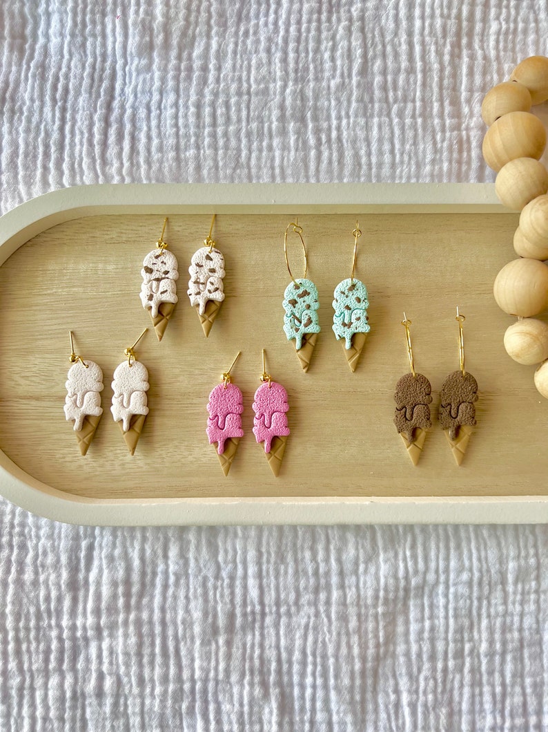 May include: A set of six pairs of ice cream cone earrings. The earrings are made of polymer clay and feature different ice cream flavors, including white, pink, mint green, and brown. The earrings are all gold-plated and have a dangling design.