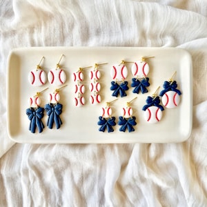 May include: A collection of baseball-themed dangle earrings featuring white circular charms with red stitching details, some accented with navy blue bows, all suspended from gold-toned metal earring hooks on a rectangular tray.