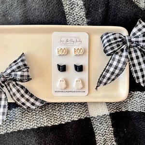 May include: A set of three pairs of stud earrings on a white display card featuring the text Seize The Clay Jewelry and seizetheclayjewelry.etsy.com. The earrings include beige BOO text, black open books, and white ghost shapes with black eyes.