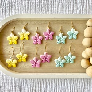 May include: Six pairs of flower-shaped earrings with gold hoops. The earrings are in three colors: yellow, pink, and light blue.