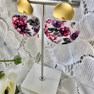 May include: A pair of white half-circle earrings with a floral pattern in shades of purple and pink. The earrings are on a gold stand.