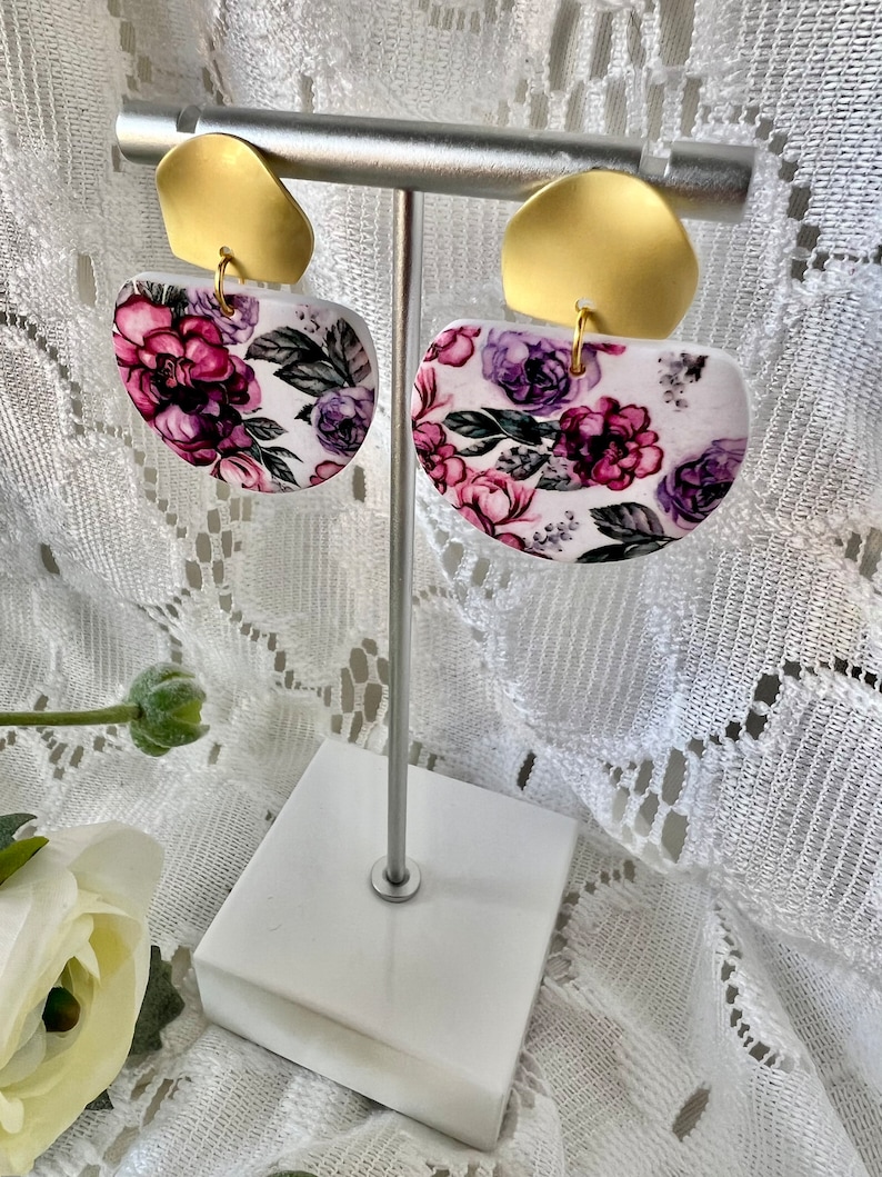 May include: A pair of white half-circle earrings with a floral pattern in shades of purple and pink. The earrings are on a gold stand.