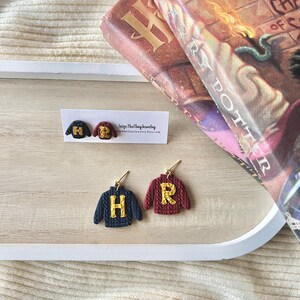 May include: Earrings shaped like miniature navy blue and burgundy sweaters, with gold-coloured letter accents. The earrings are displayed on a white tray, with a book in the background. The letters "H" and "R" are visible on the sweaters.
