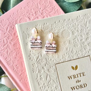 Peut inclure: Une paire de boucles d'oreilles en forme de livre rose et blanc avec un accent de perle. Les boucles d'oreilles sont sur un fond blanc avec le texte "Write the Word" et "The Fruit of the Spirit".
