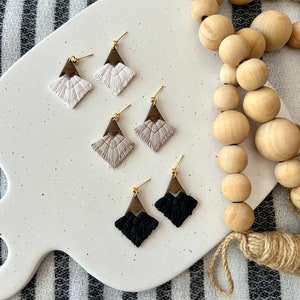 May include: Three pairs of earrings with a geometric design. The earrings are made of wood and have a fringe detail. The earrings are in white, beige, and black.