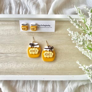 May include: A pair of honey jar earrings with a white background. The earrings are made of polymer clay and have a gold finish. The jars are decorated with the word "honey" in brown lettering. The earrings are on a white card with the text "Seize The Clay Jewelry" and a website address.