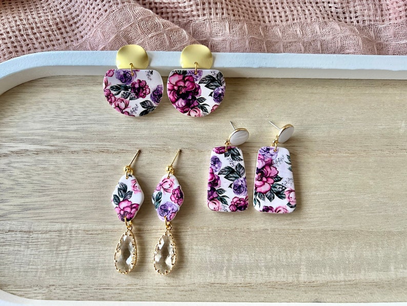 May include: Three pairs of floral print earrings. The first pair is a semi-circle shape with gold accents. The second pair is a rectangle shape with gold accents. The third pair is a teardrop shape with a clear crystal and gold accents.