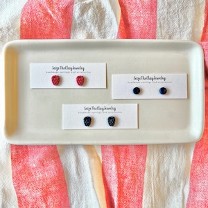 May include: A white rectangular tray displays three pairs of earrings. One pair is red raspberry-shaped, another is dark blue studs, and the third is black blackberry-shaped. Each pair is on a card with the text "Seize The Clay Jewelry handmade earrings and accessories."