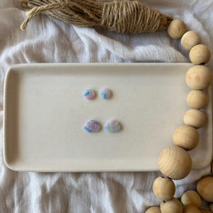 May include: Two pairs of small stud earrings featuring a marbled pattern in shades of light blue, pink, and white. One pair is circular in shape, while the other pair is shaped like small clouds. The earrings are displayed on a rectangular tray.