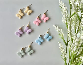 May include: A set of six pairs of butterfly earrings in pastel colours, including yellow, pink, lavender, and blue. The earrings are displayed on a white plate with a scalloped edge.