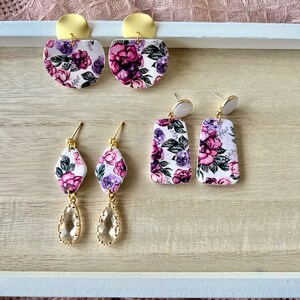 May include: A set of three pairs of floral earrings. The first pair features a large, round, white base with a purple and green floral pattern and gold accents. The second pair features a smaller, rectangular, white base with a purple and green floral pattern. The third pair features a teardrop-shaped, clear crystal with gold accents.