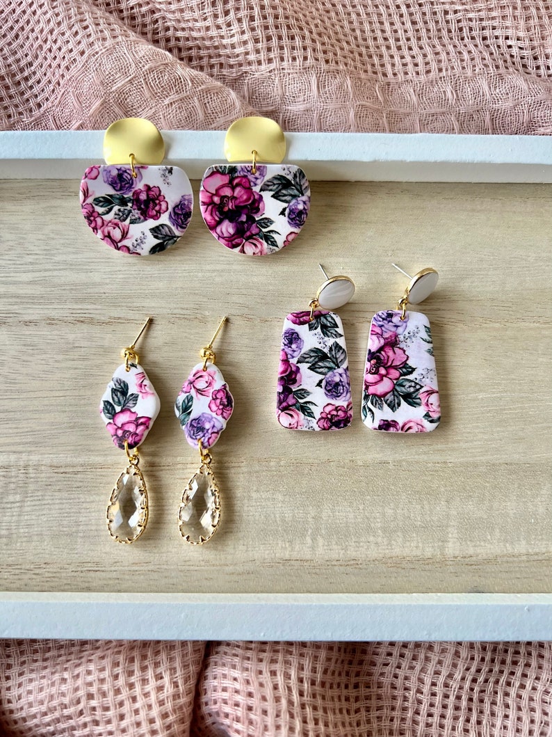 May include: A set of three pairs of floral earrings. The first pair features a large, round, white base with a purple and green floral pattern and gold accents. The second pair features a smaller, rectangular, white base with a purple and green floral pattern. The third pair features a teardrop-shaped, clear crystal with gold accents.