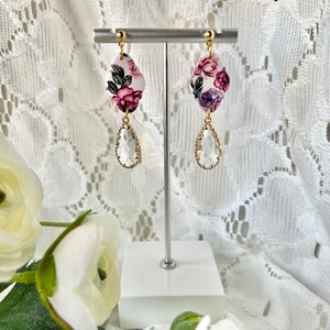 May include: A pair of floral dangle earrings with a white background and pink, purple, and green flowers. The earrings have a clear teardrop-shaped crystal hanging from the floral charm.
