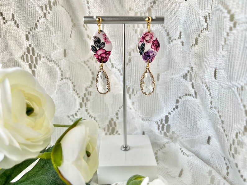 May include: A pair of floral dangle earrings with a white background and pink, purple, and green flowers. The earrings have a clear teardrop-shaped crystal hanging from the floral charm.