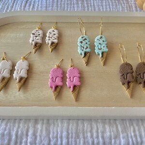 May include: A set of six pairs of ice cream cone earrings. The earrings are made of polymer clay and come in four different colors: white, pink, blue, and brown. Each pair of earrings features a double scoop of ice cream with a chocolate drizzle.