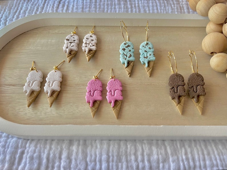 May include: A set of six pairs of ice cream cone earrings. The earrings are made of polymer clay and come in four different colors: white, pink, blue, and brown. Each pair of earrings features a double scoop of ice cream with a chocolate drizzle.