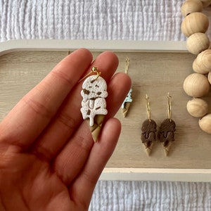 May include: A pair of ice cream cone earrings with a white and brown ice cream design. The earrings are made of polymer clay and have gold hooks.