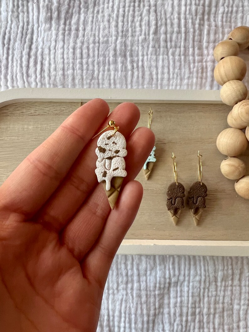 May include: A pair of ice cream cone earrings with a white and brown ice cream design. The earrings are made of polymer clay and have gold hooks.