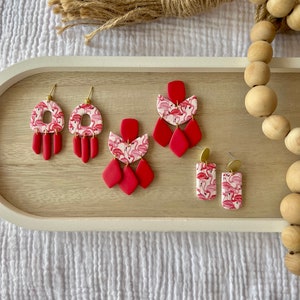 May include: A set of four pairs of pink flamingo earrings. The earrings are made of polymer clay and have gold accents. The earrings are displayed on a wooden tray.