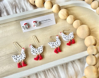 May include: A set of three pairs of chicken earrings. The earrings are made of white polymer clay with black polka dots and red boots. The earrings are displayed on a wooden tray with a wooden bead garland.