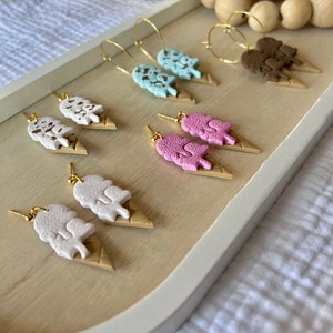 May include: A set of six pairs of ice cream cone earrings. The earrings are made of polymer clay and feature different ice cream flavors, including chocolate, vanilla, strawberry, and mint chocolate chip. The earrings are all attached to gold hoops.