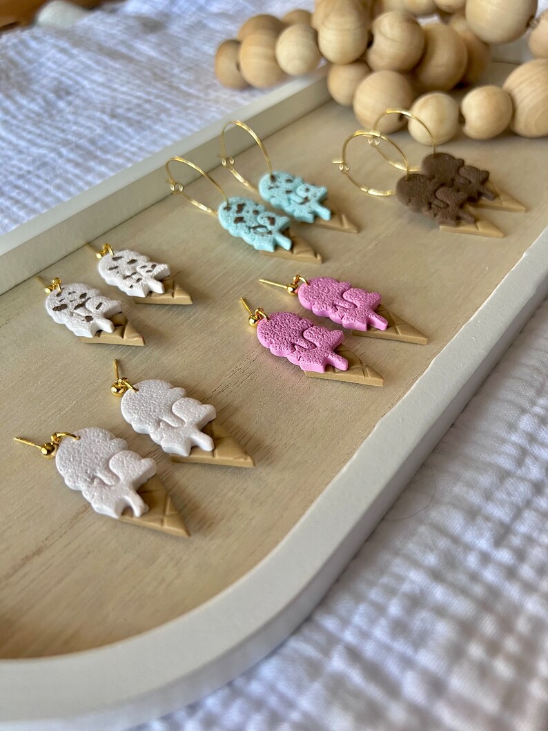 May include: A set of six pairs of ice cream cone earrings. The earrings are made of polymer clay and feature different ice cream flavors, including chocolate, vanilla, strawberry, and mint chocolate chip. The earrings are all attached to gold hoops.
